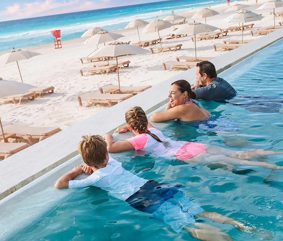 Family on vacation in an infinity pool overlooking the beach with chairs and beach umbrellas.